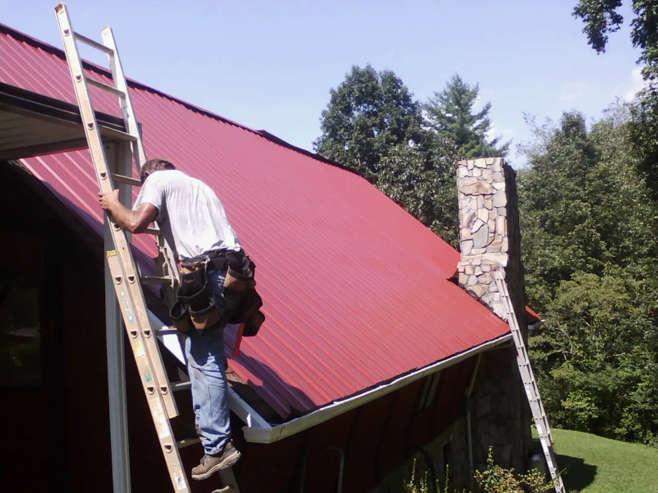 Roofer on a ladder beside a red metal roof and stone chimney — Soffit & Fascia Repair by Roofing Brothers in Reed Point